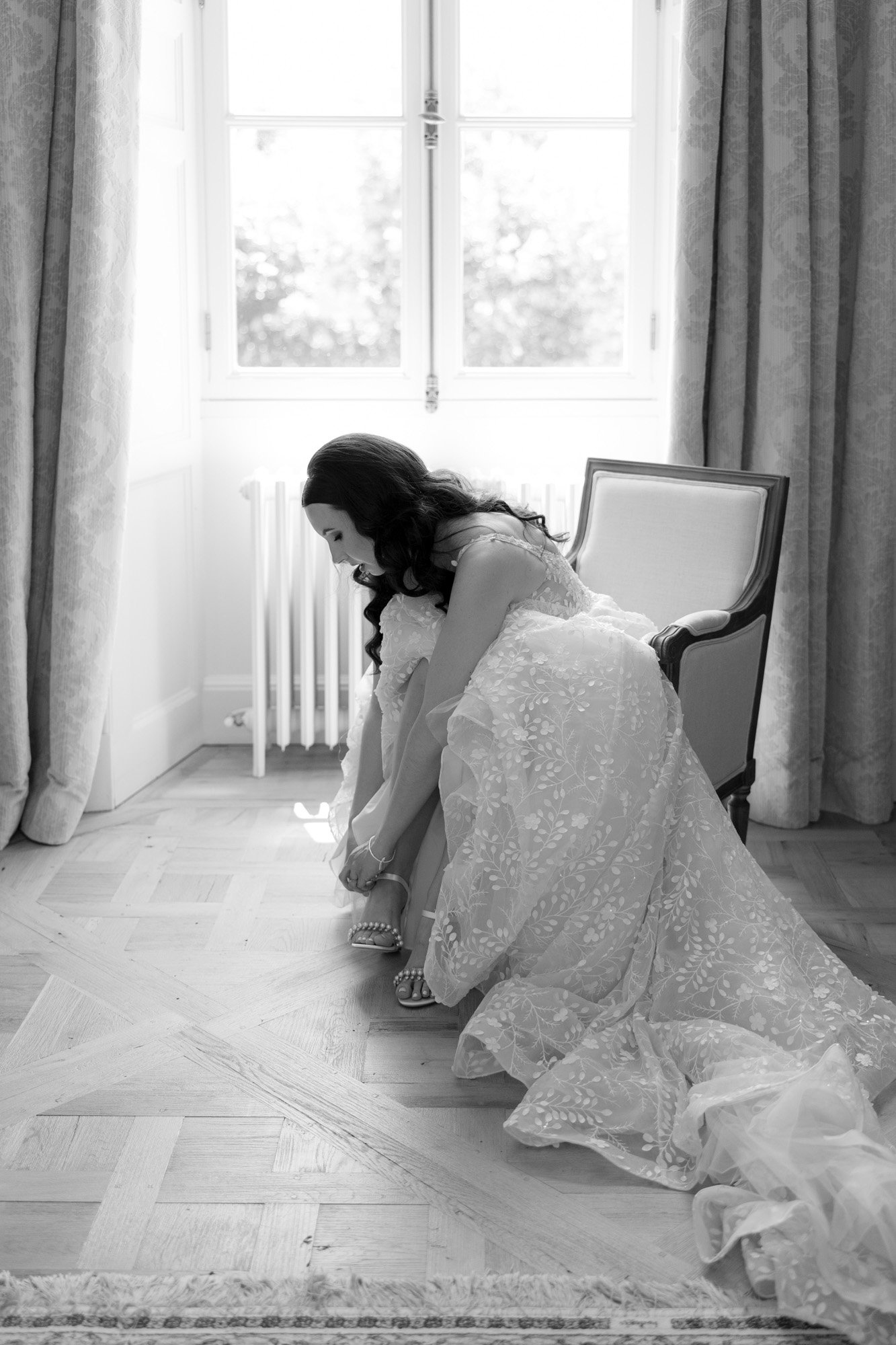 A woman in a wedding dress sits on a chair and adjusts her shoe in a bright room with a window and curtains.