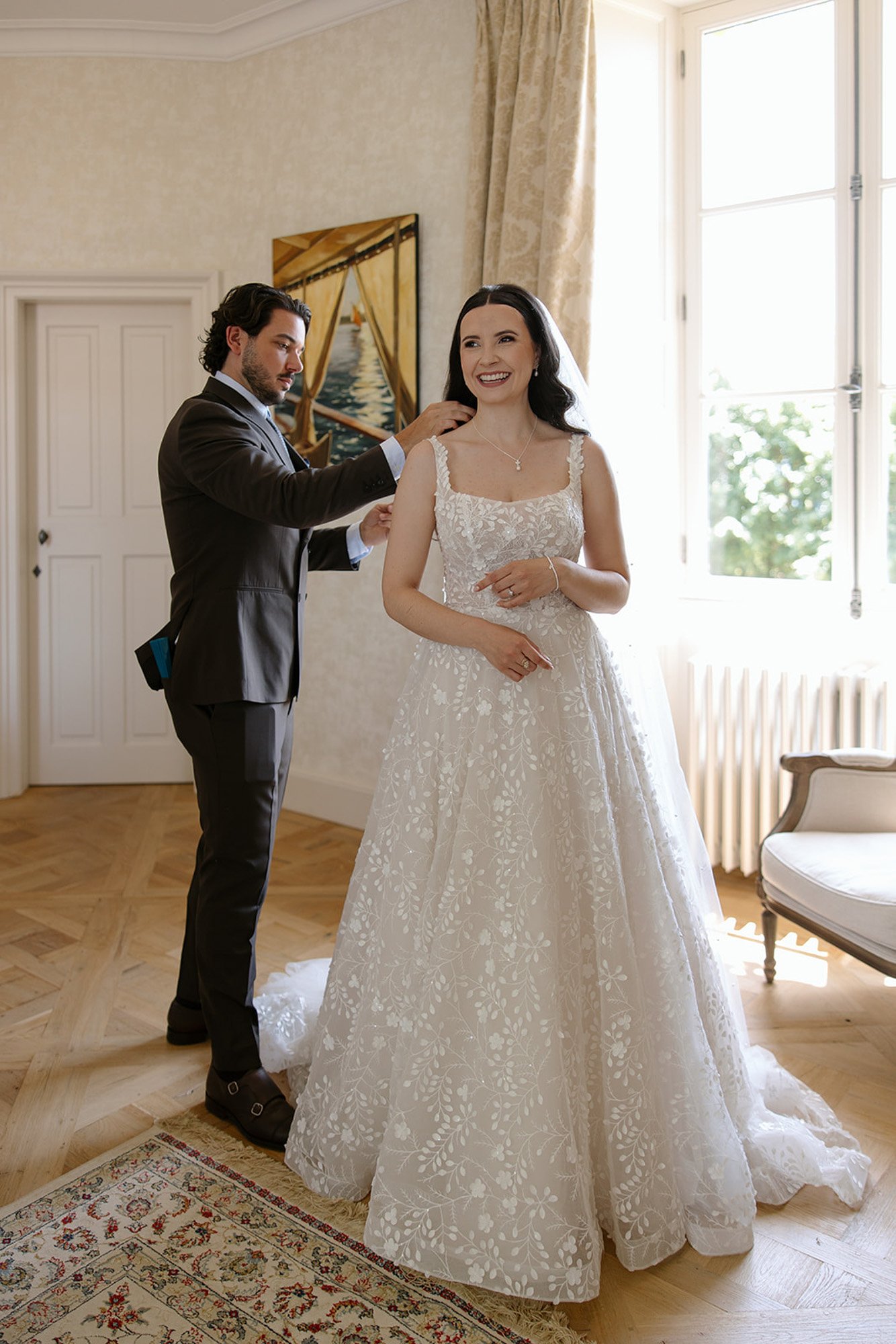 A bride in a white floral wedding dress smiles while a man in a suit stands behind her, adjusting her hair in a bright room.
