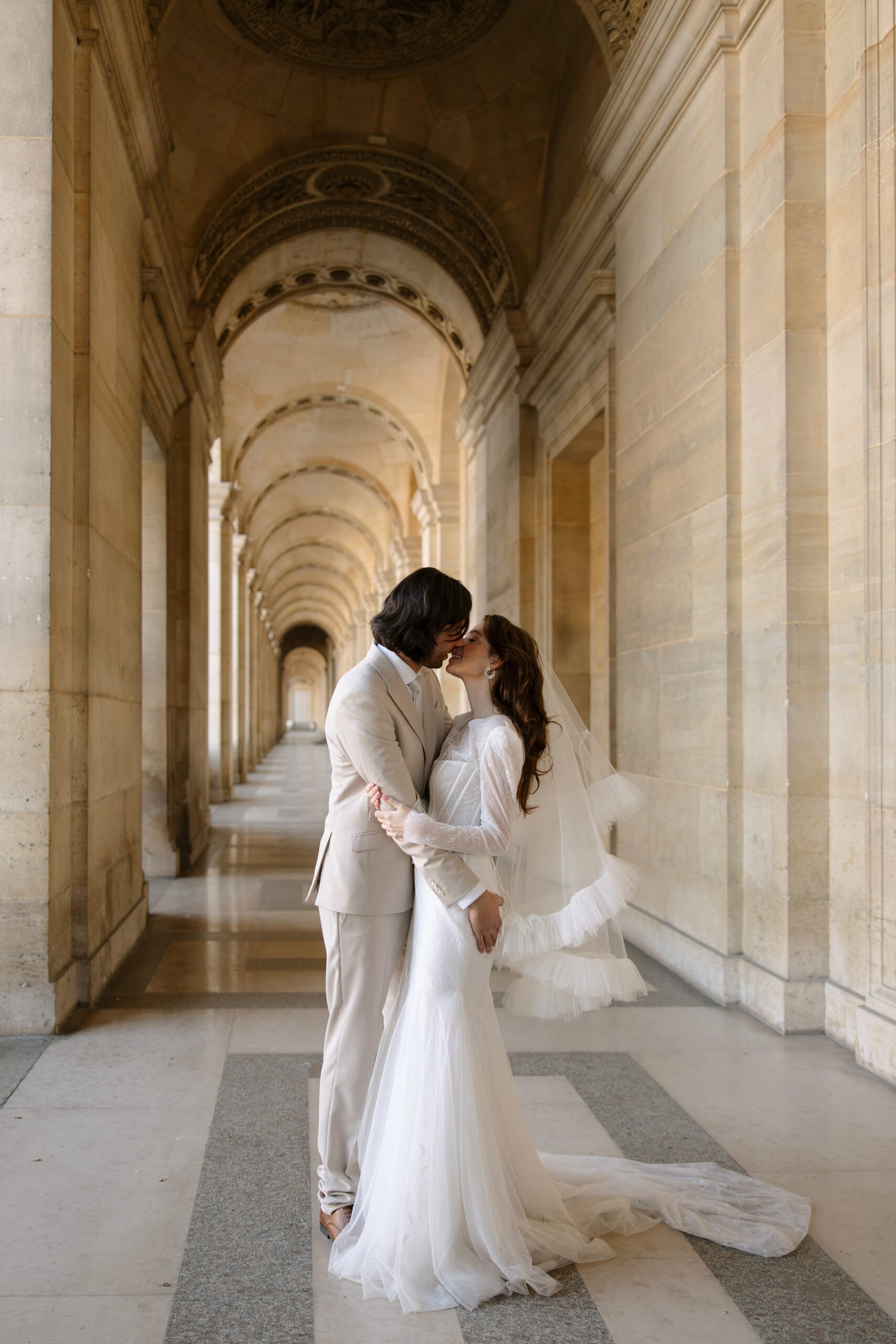 A bride and groom, dressed in a white dress and light suit, kiss at the Louvre in Paris in an ornate, arched corridor with stone columns and a high, decorated ceiling. Captured by Paris wedding photographer.