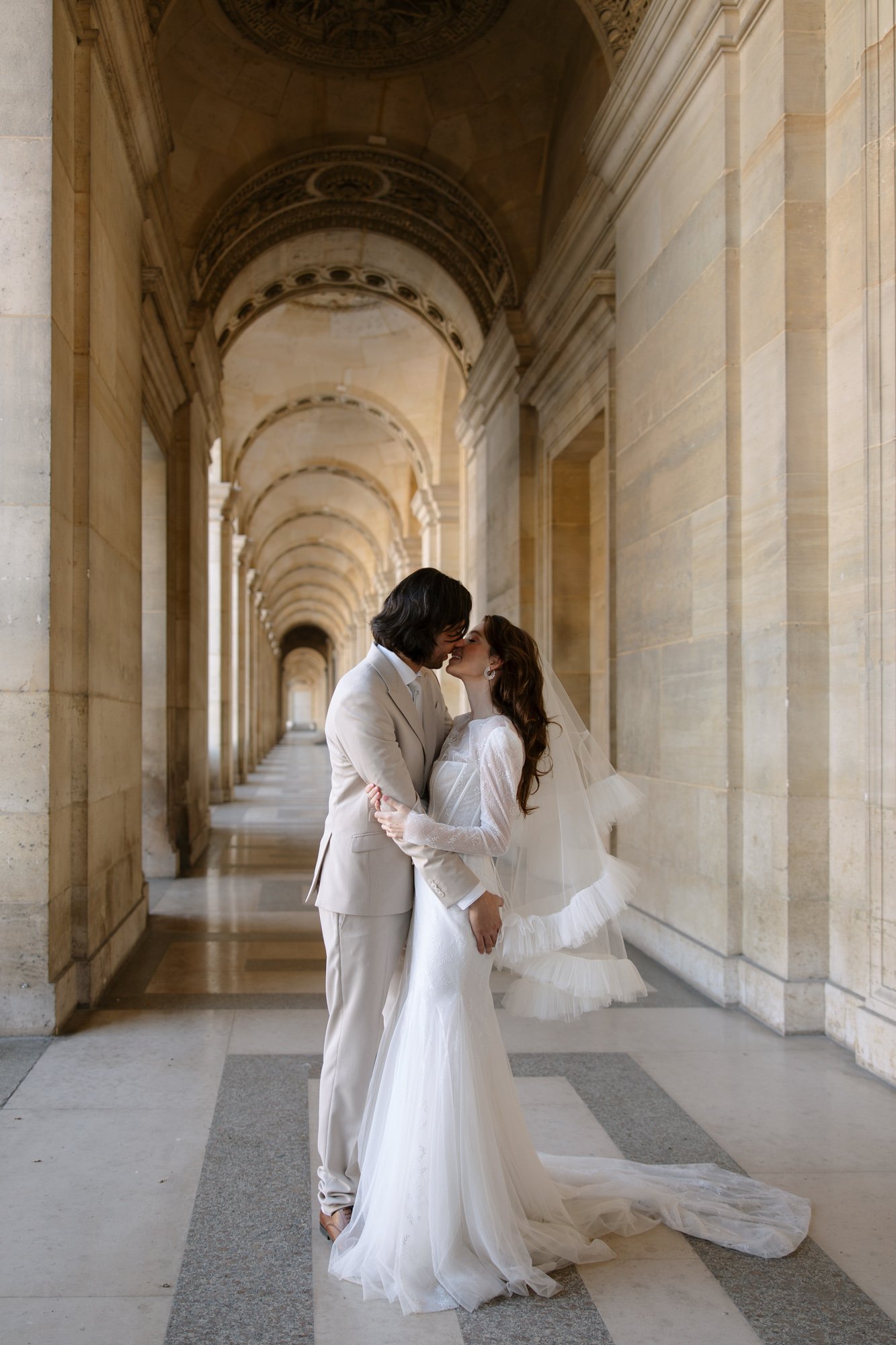 A bride and groom kiss under a grand stone archway with ornate ceilings and columns, dressed in formal wedding attire. Paris wedding photographer.