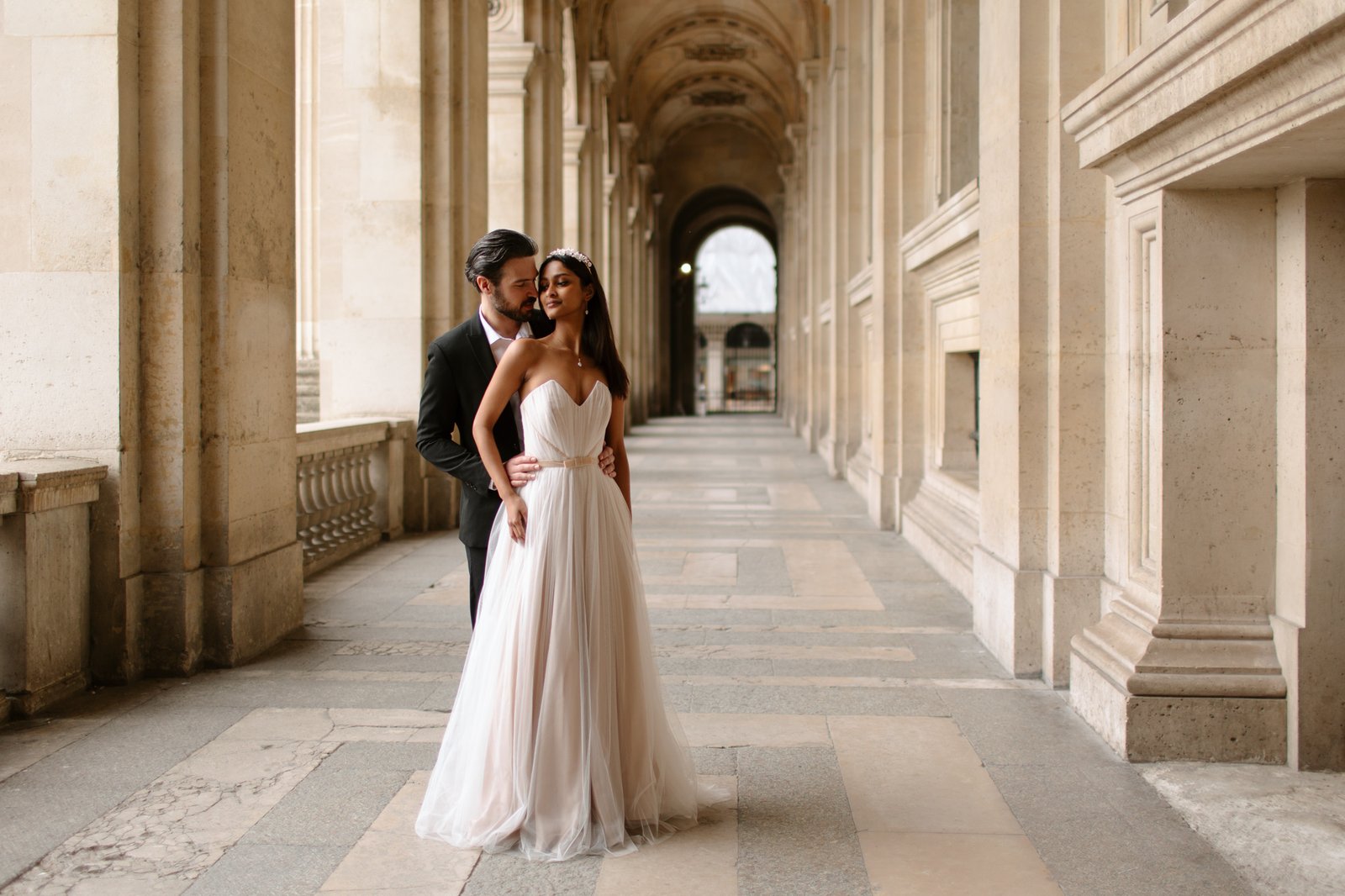 A bride in a strapless white gown and a groom in a dark suit stand close together in an elegant, arched stone corridor. Paris Wedding Photoshoot