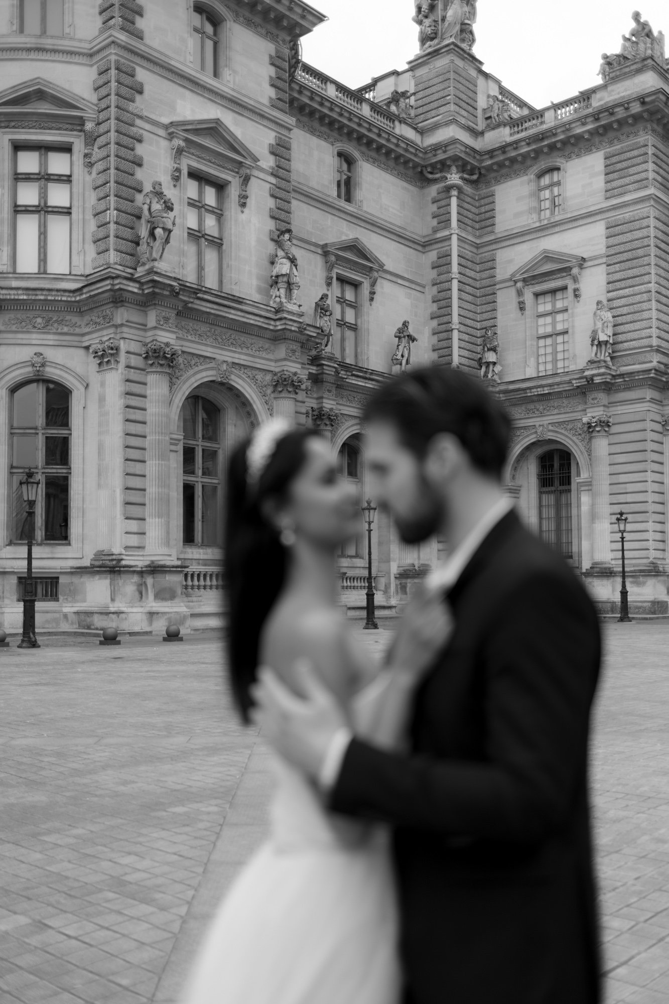 A bride and groom stand closely together in focus, with a historic stone building featuring large windows and statues in the background.