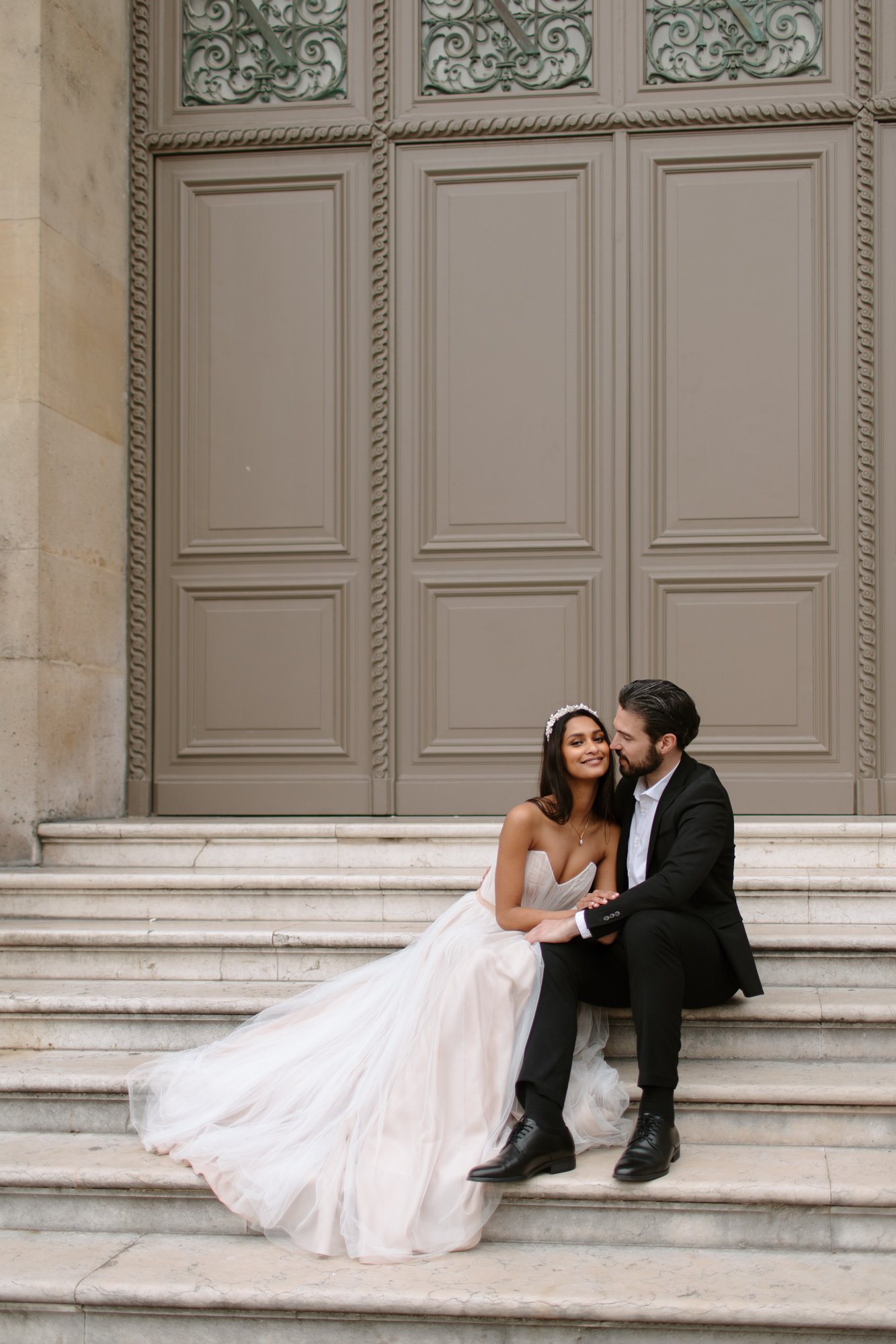 A bride in a white gown and a groom in a black suit sit together on stone steps in front of large, ornate doors, smiling at each other. Paris Wedding Photoshoot