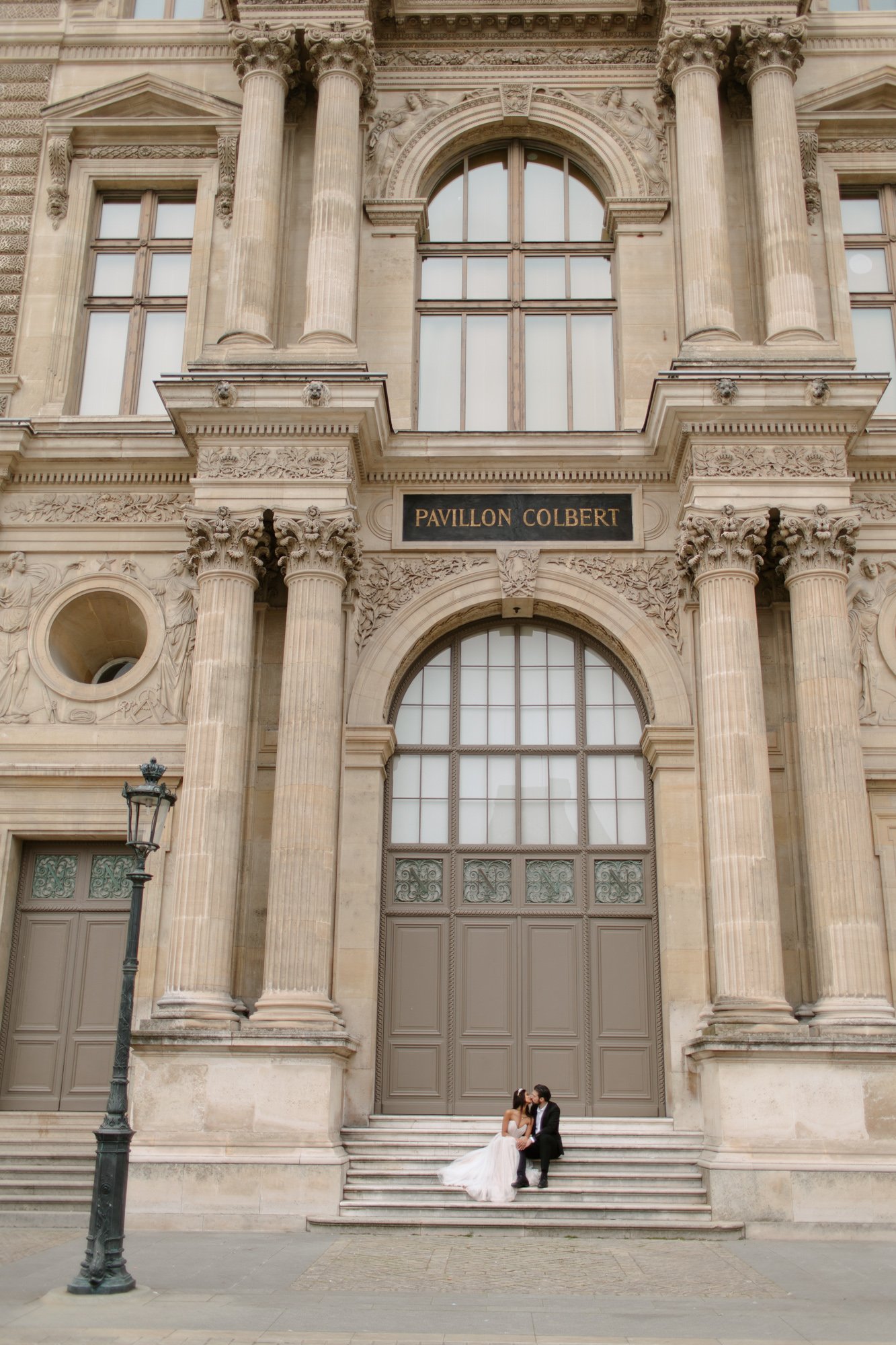 A bride and groom sit on the steps in front of the Pavillon Colbert, a grand building with tall arched windows and ornate stone columns. Paris Wedding Photoshoot
