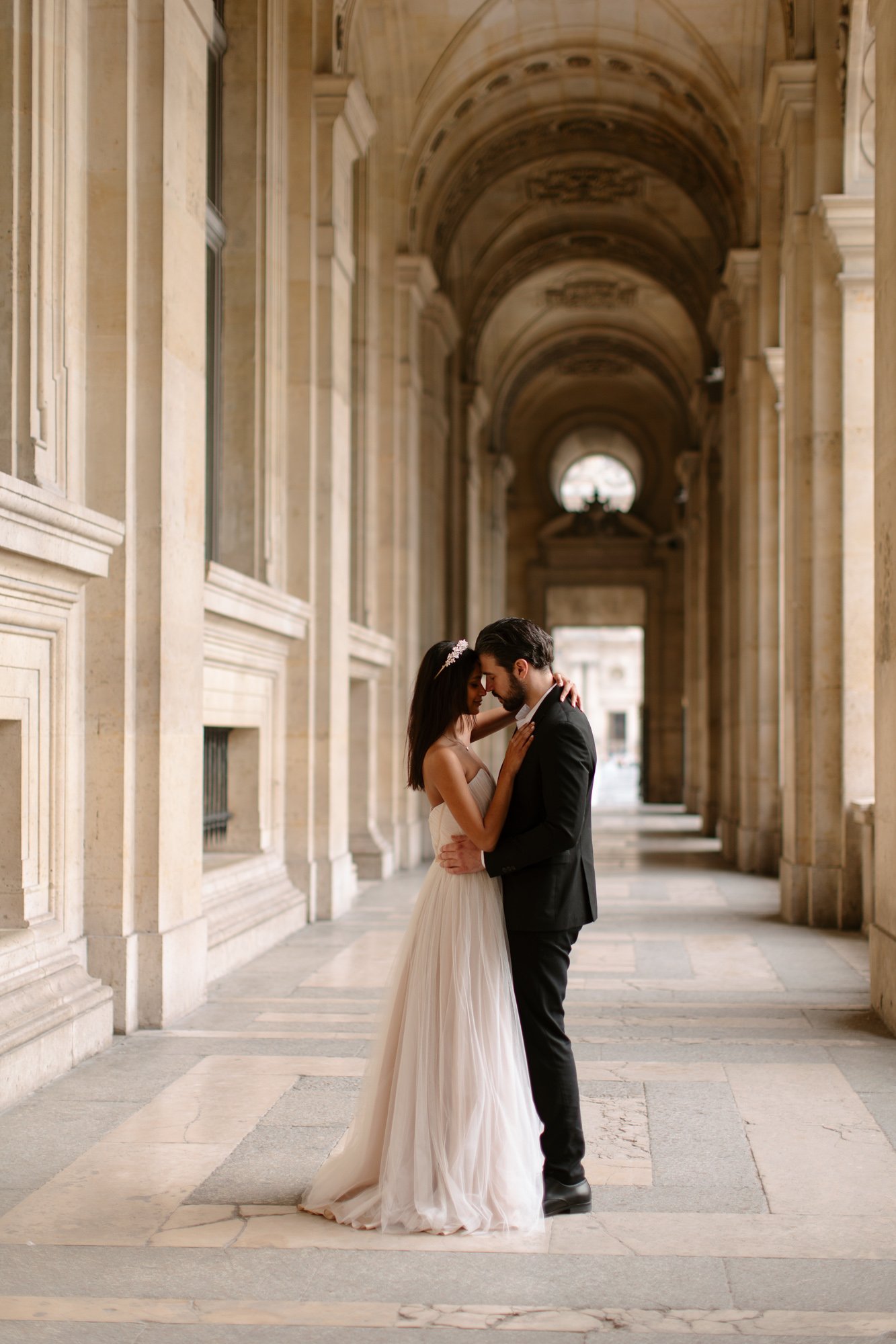 A couple dressed in formal attire, with the woman in a white gown and the man in a black suit, embrace in an ornate, arched hallway. Paris Wedding Photoshoot