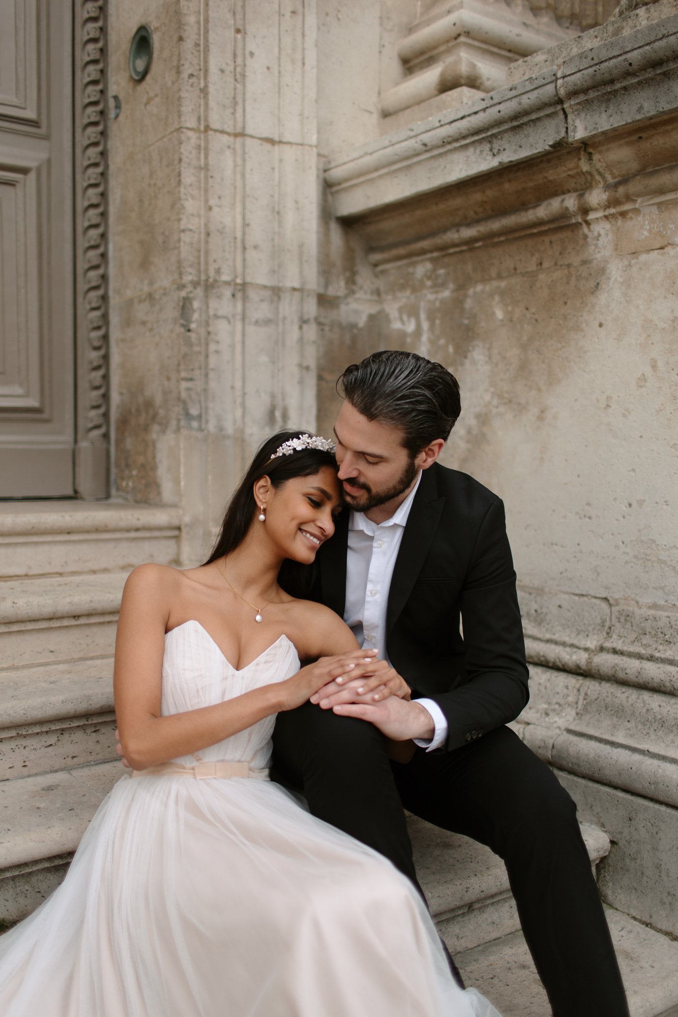 A bride in a white gown and a groom in a black suit sit on stone steps, holding hands and smiling affectionately at each other.