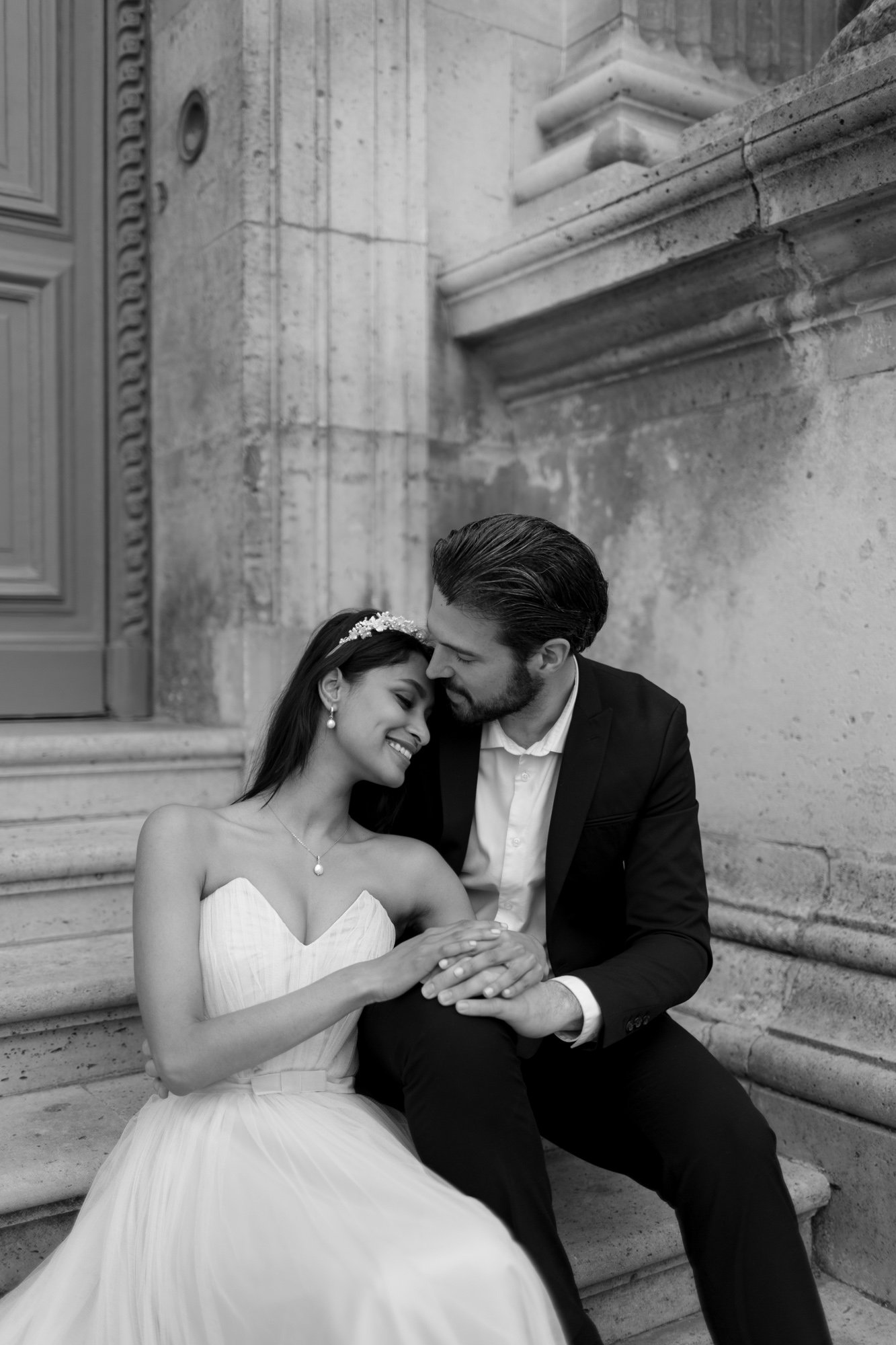 A bride and groom sit closely together on stone steps, holding hands and smiling at each other, dressed in wedding attire. Paris Wedding Photoshoot