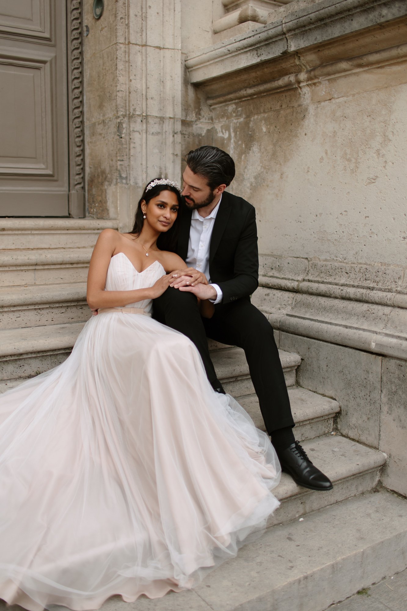 A bride in a white gown and a groom in a black suit sit closely together on stone steps outside a building, holding hands and looking at each other.