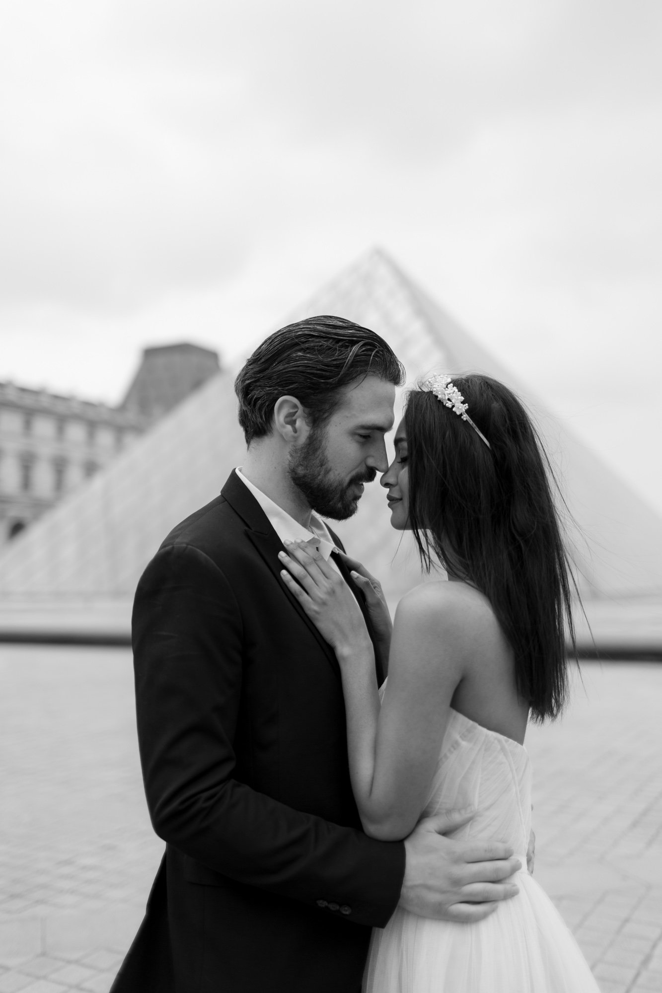 A couple in formal attire stands closely together in front of the Louvre Pyramid, with their foreheads touching.