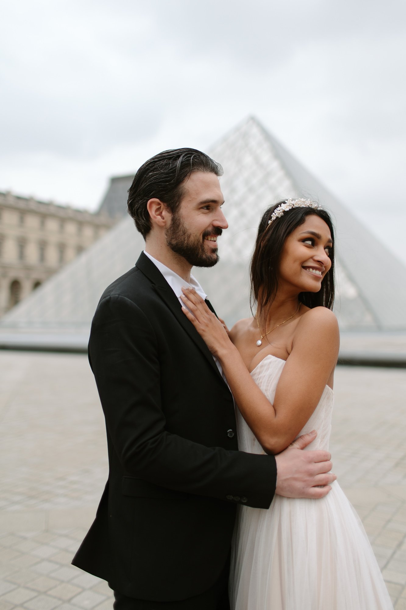 A couple in wedding attire poses in front of the Louvre Pyramid in Paris, with the bride smiling and the groom looking ahead. Paris Wedding Photoshoot