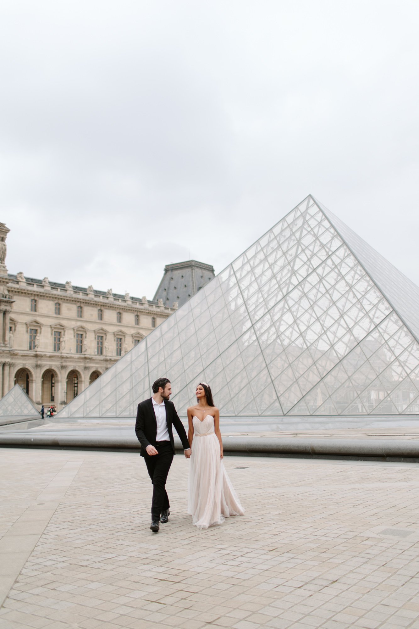 A couple in formal attire walks hand in hand in front of the glass pyramid at the Louvre Museum in Paris on a cloudy day. Paris Wedding Photoshoot