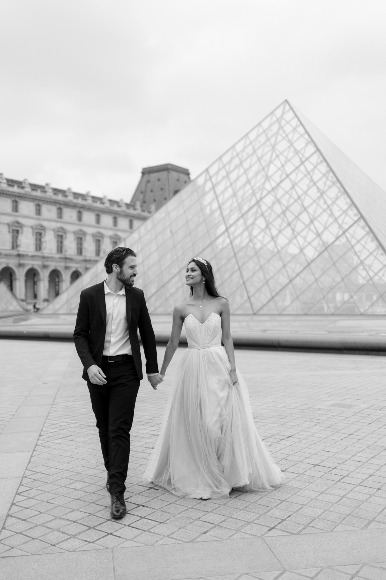 A couple dressed in formal wedding attire walks hand in hand outside the Louvre Pyramid in Paris.