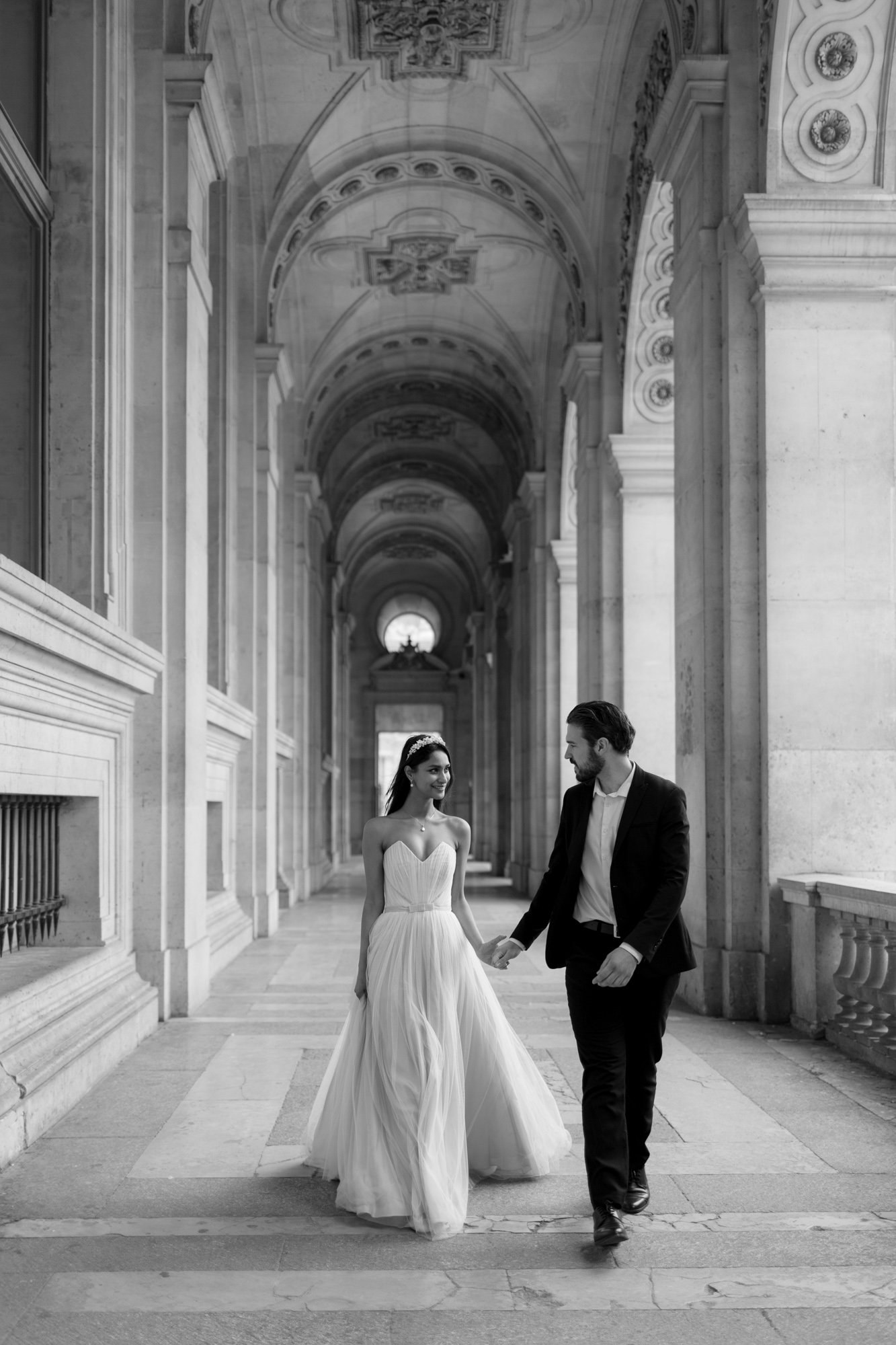 A bride in a strapless gown and a groom in a suit walk hand in hand through an ornate, arched hallway. Paris Wedding Photoshoot