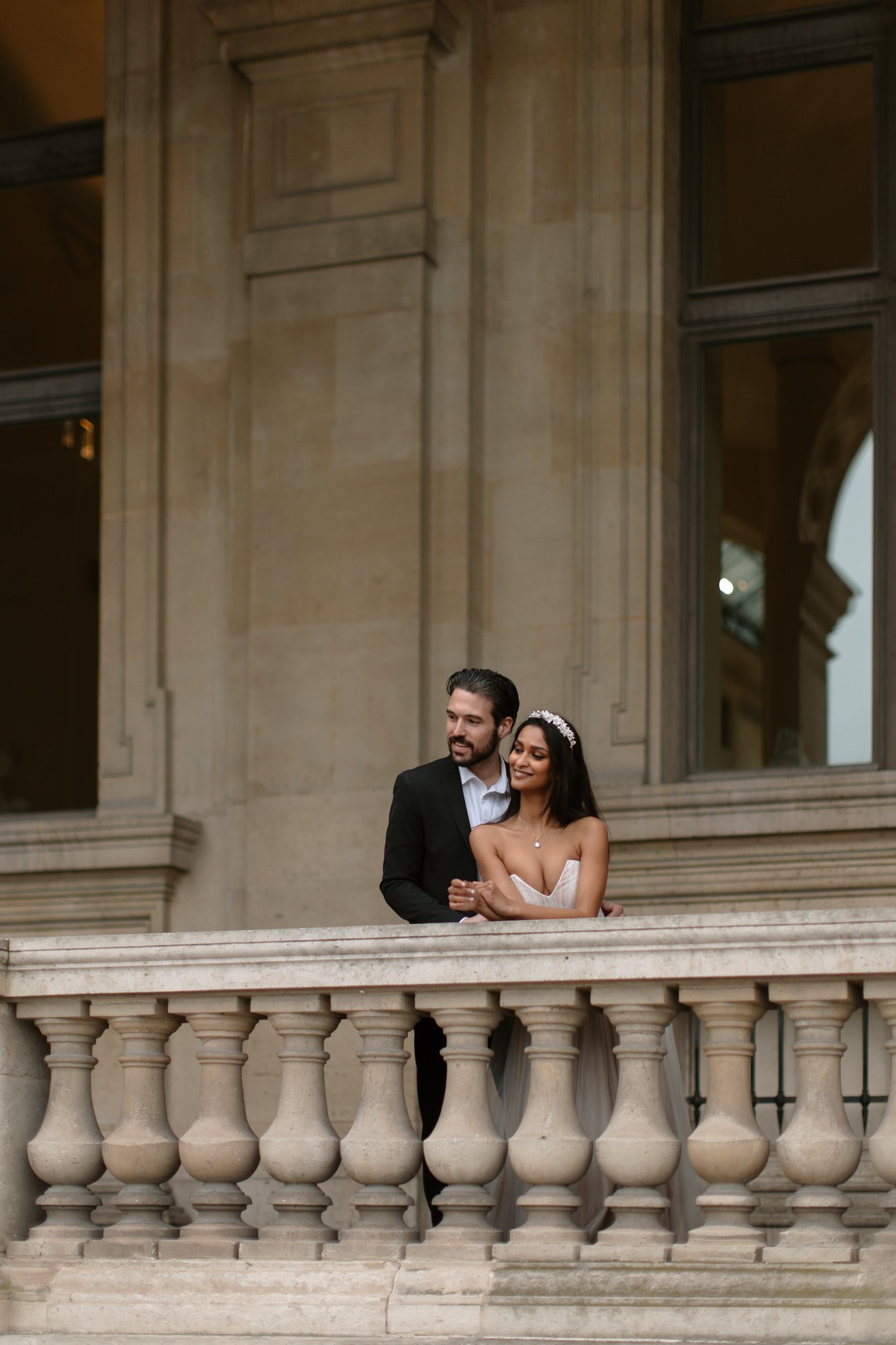 A man in a suit and a woman in a strapless dress stand together on a stone balcony, posing in front of a large building with tall windows. Paris Wedding Photoshoot