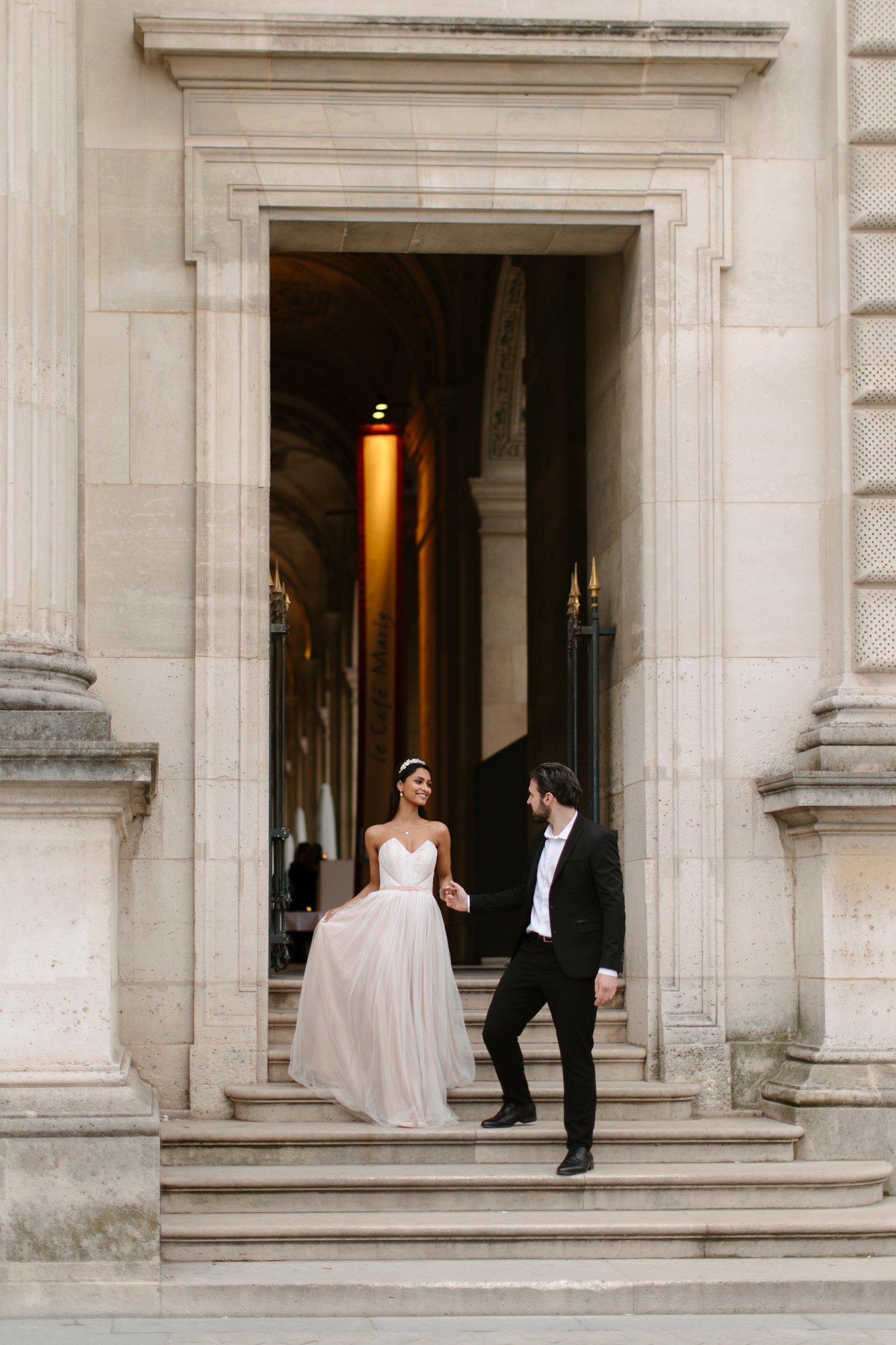 A woman in a white gown and a man in a black suit stand on stone steps outside a grand building, holding hands and looking at each other.