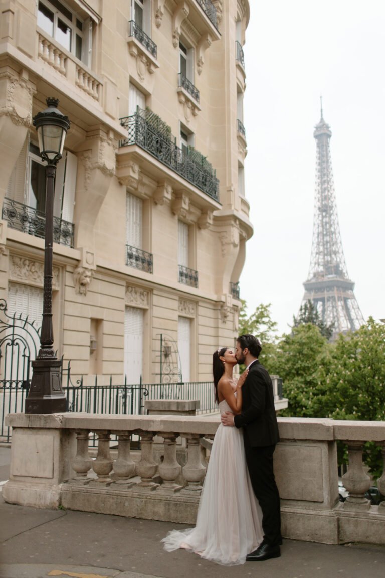 A couple in wedding attire stands embracing on a Paris street near a stone railing, with the Eiffel Tower visible in the background.