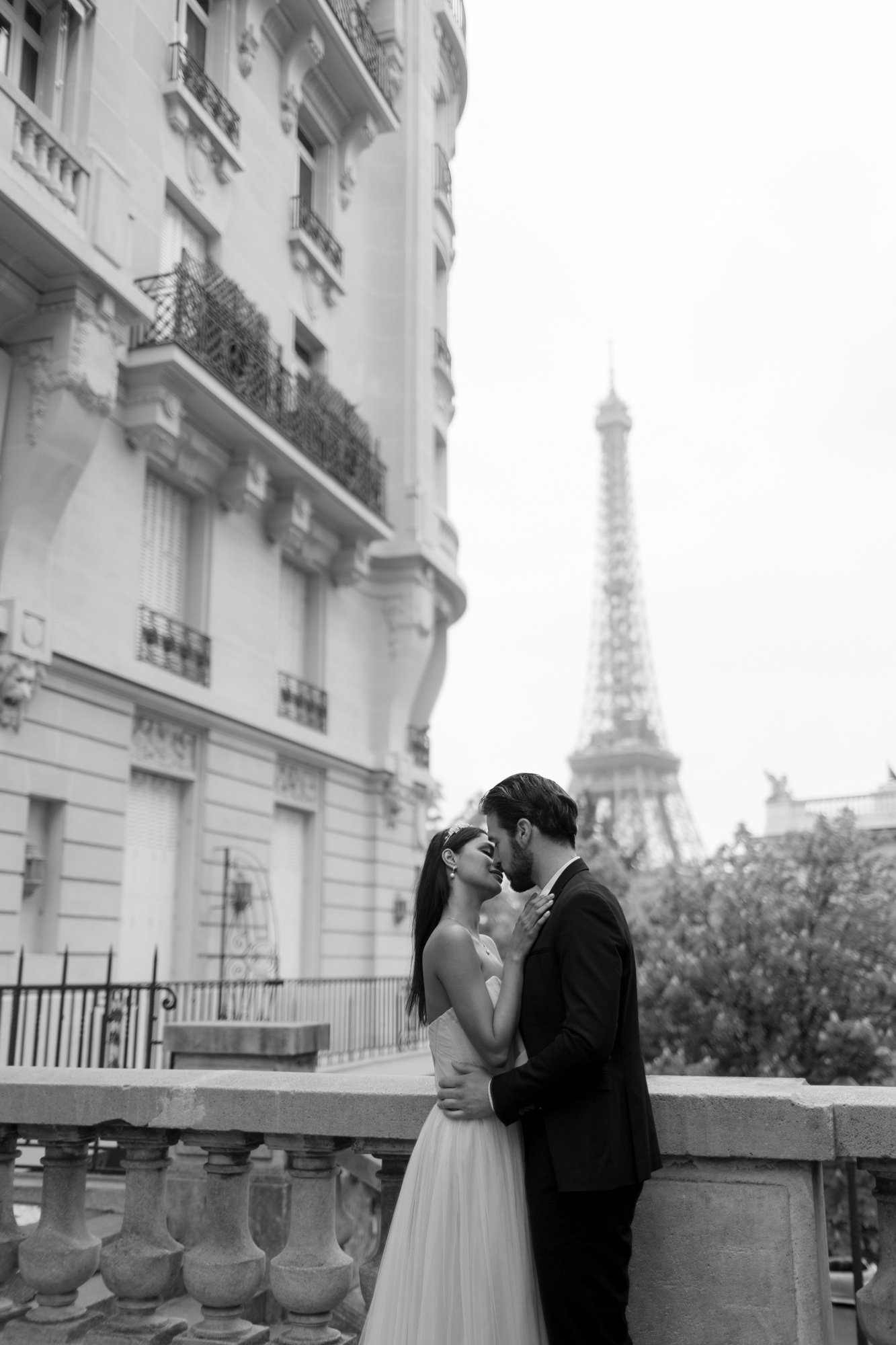 A couple dressed formally embraces on a balcony with the Eiffel Tower visible in the background; buildings and trees surround them. Paris Wedding Photoshoot