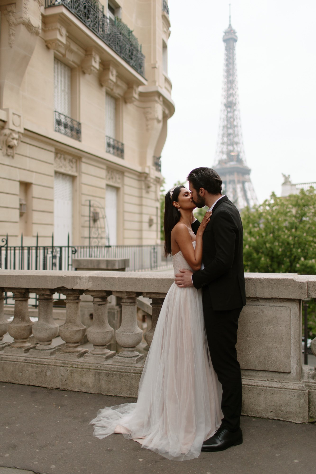 A bride and groom kiss on a Parisian street with the Eiffel Tower visible in the background.