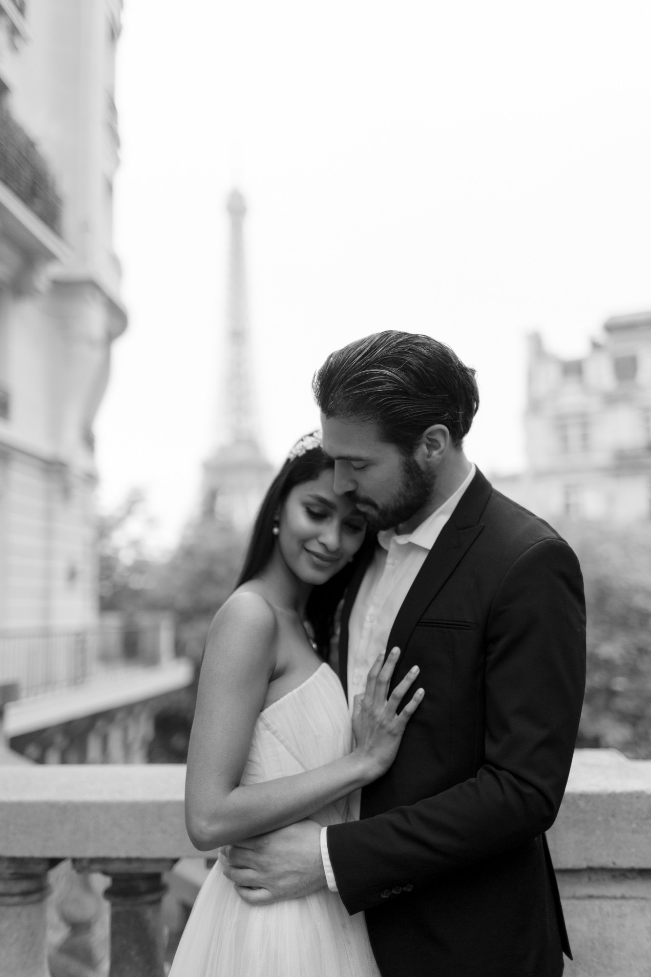 A couple embraces on a balcony with the Eiffel Tower in the background. The scene is in black and white. 