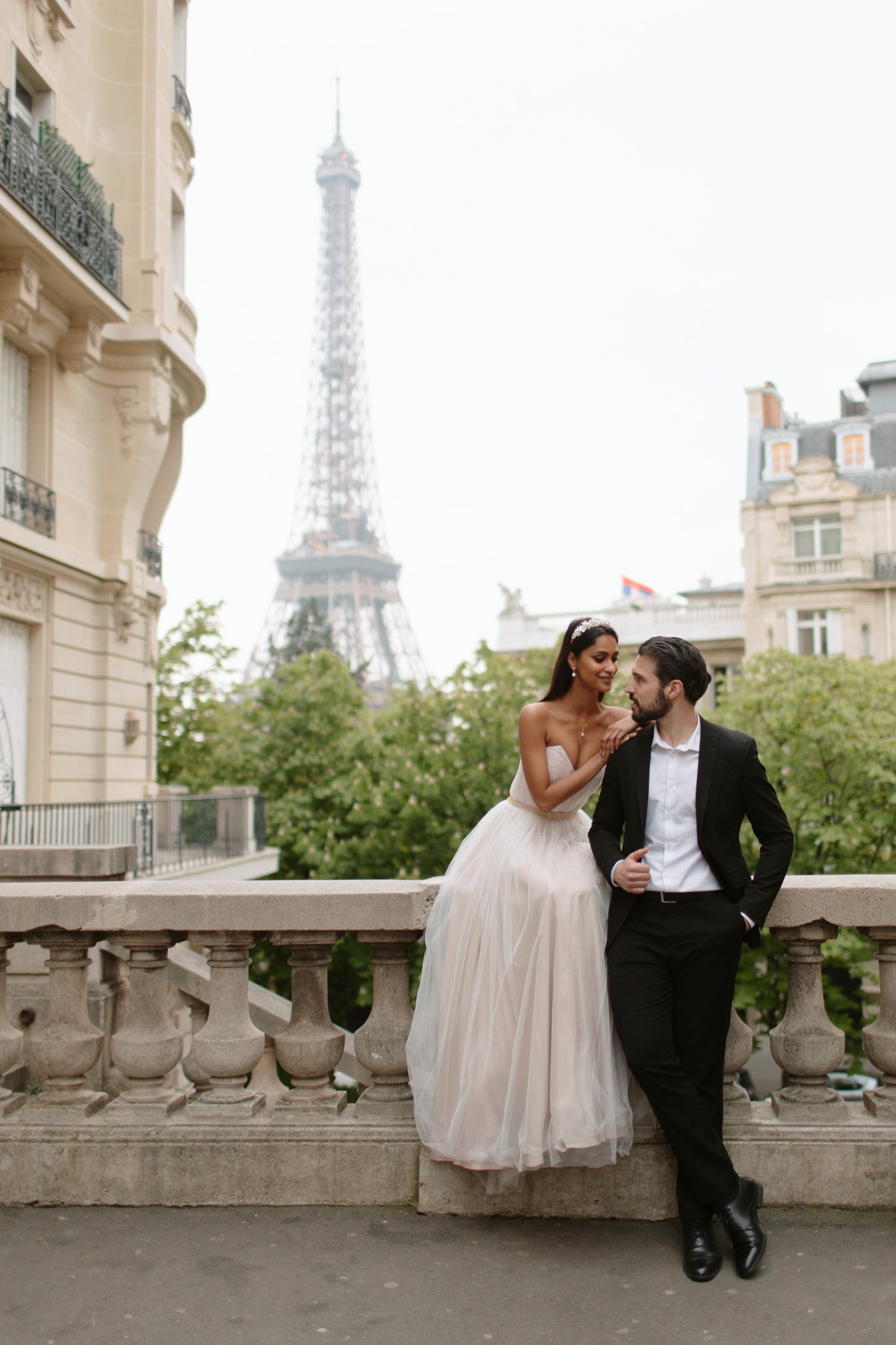 A bride and groom pose together on a balcony with the Eiffel Tower visible in the background, surrounded by Parisian buildings and greenery. 