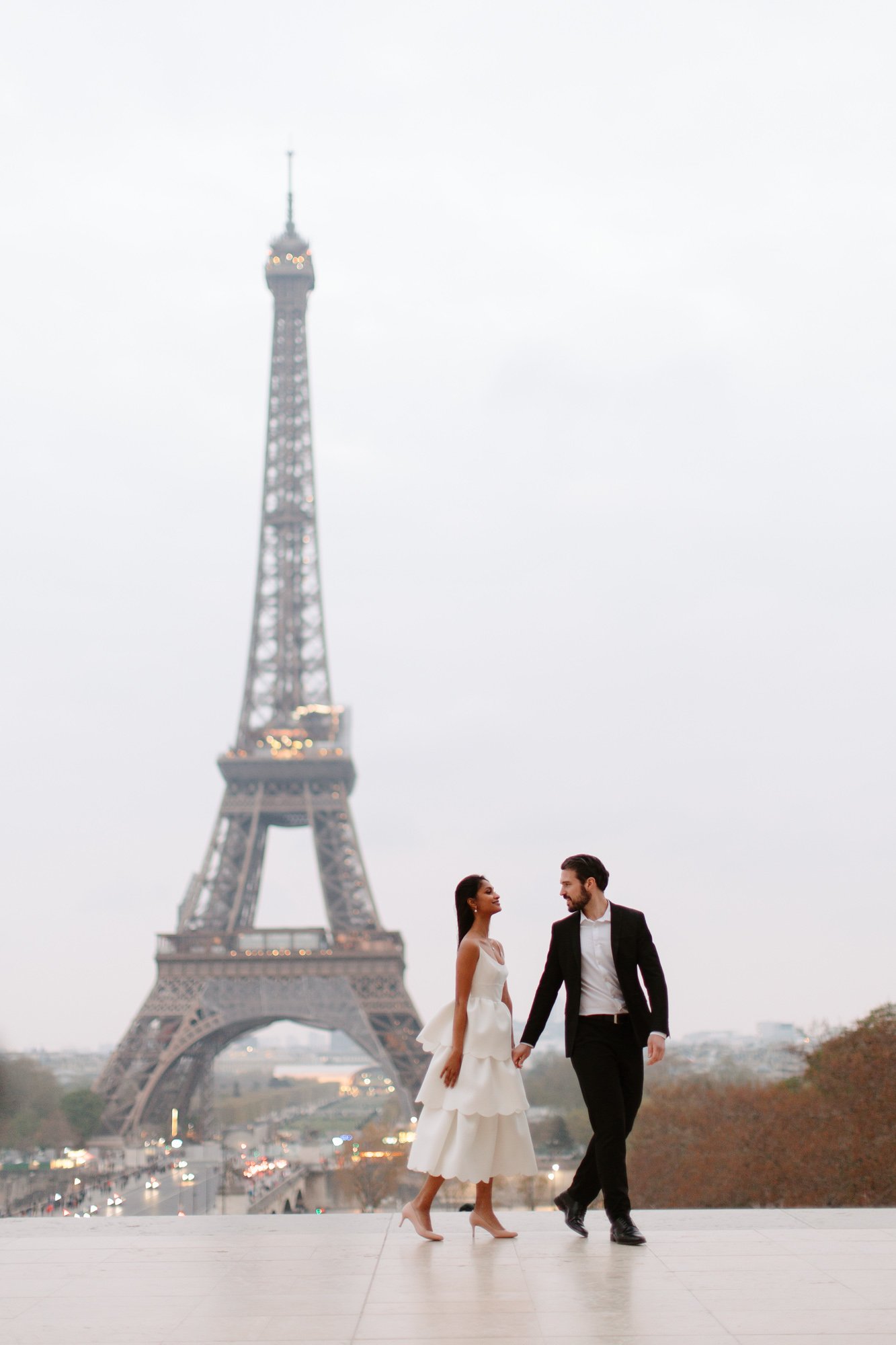 A couple in formal attire walks hand in hand near the Eiffel Tower in Paris on a cloudy day.