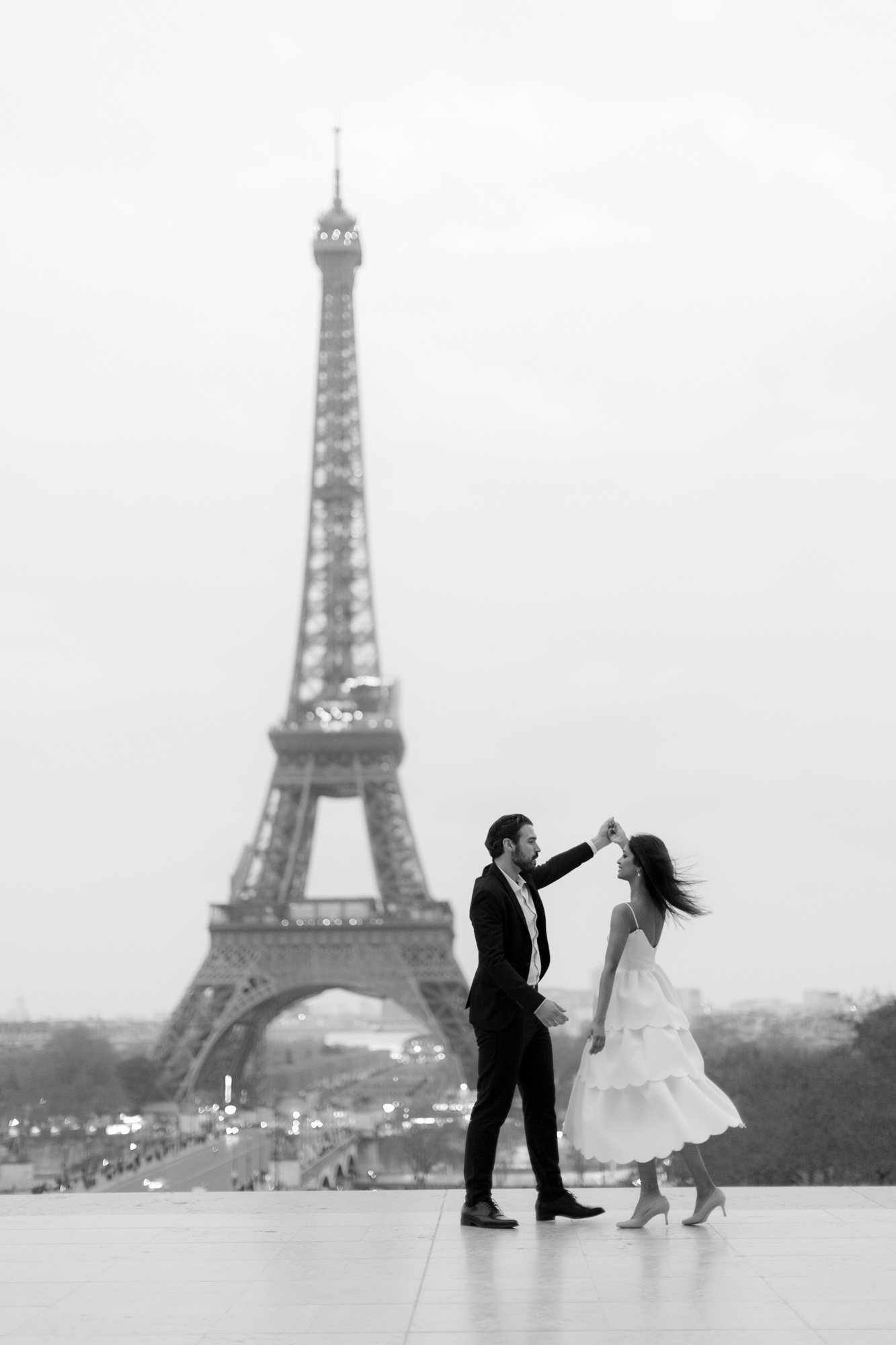 A couple dances in front of the Eiffel Tower in Paris on a cloudy day. The image is in black and white. 