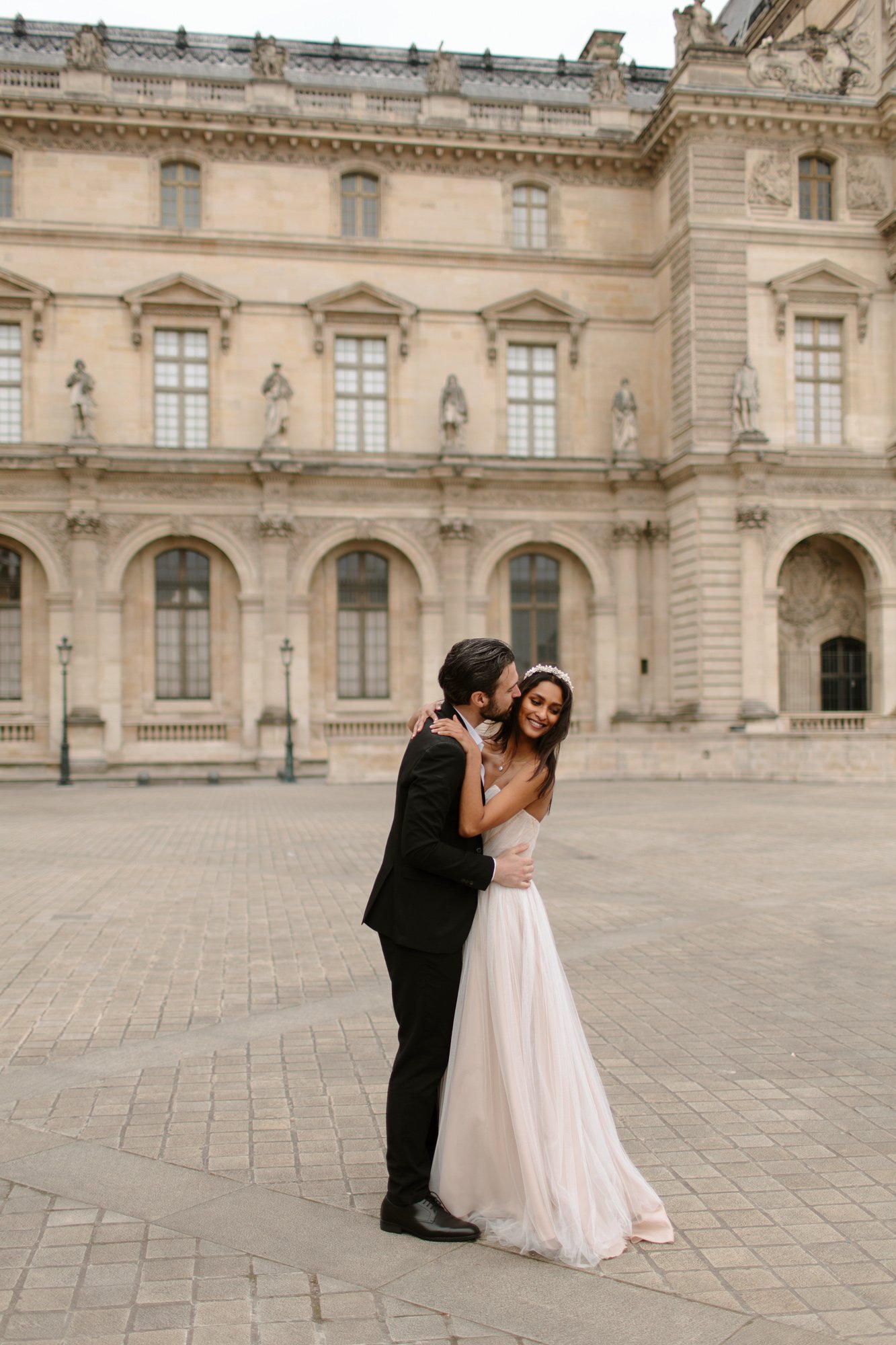 A couple dressed in formal wedding attire embraces and smiles outside a historic stone building with arched windows and statues. Paris Wedding Photoshoot