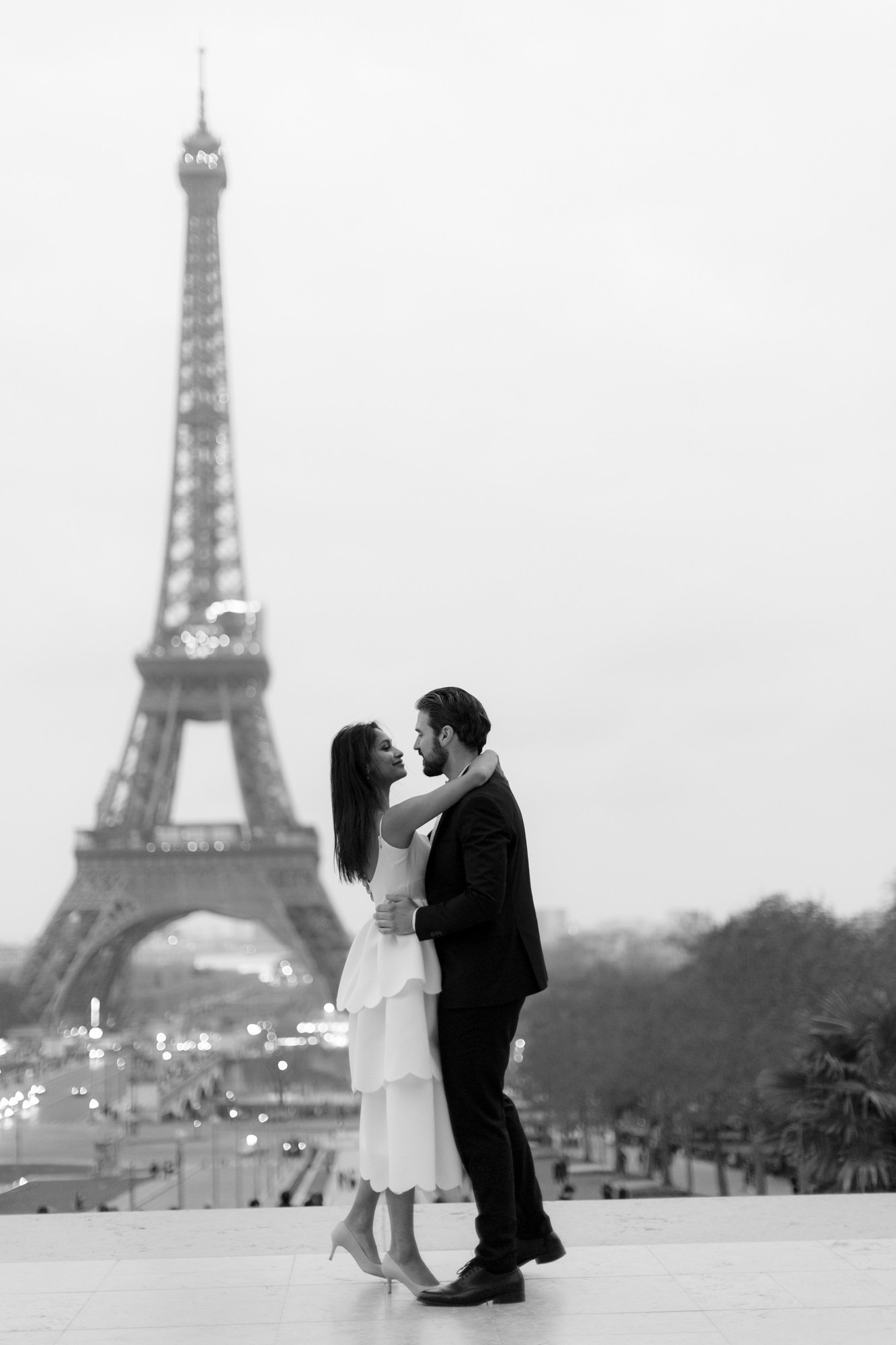 A couple dressed formally embraces in front of the Eiffel Tower in Paris, with the scene captured in black and white. Paris wedding photographer.