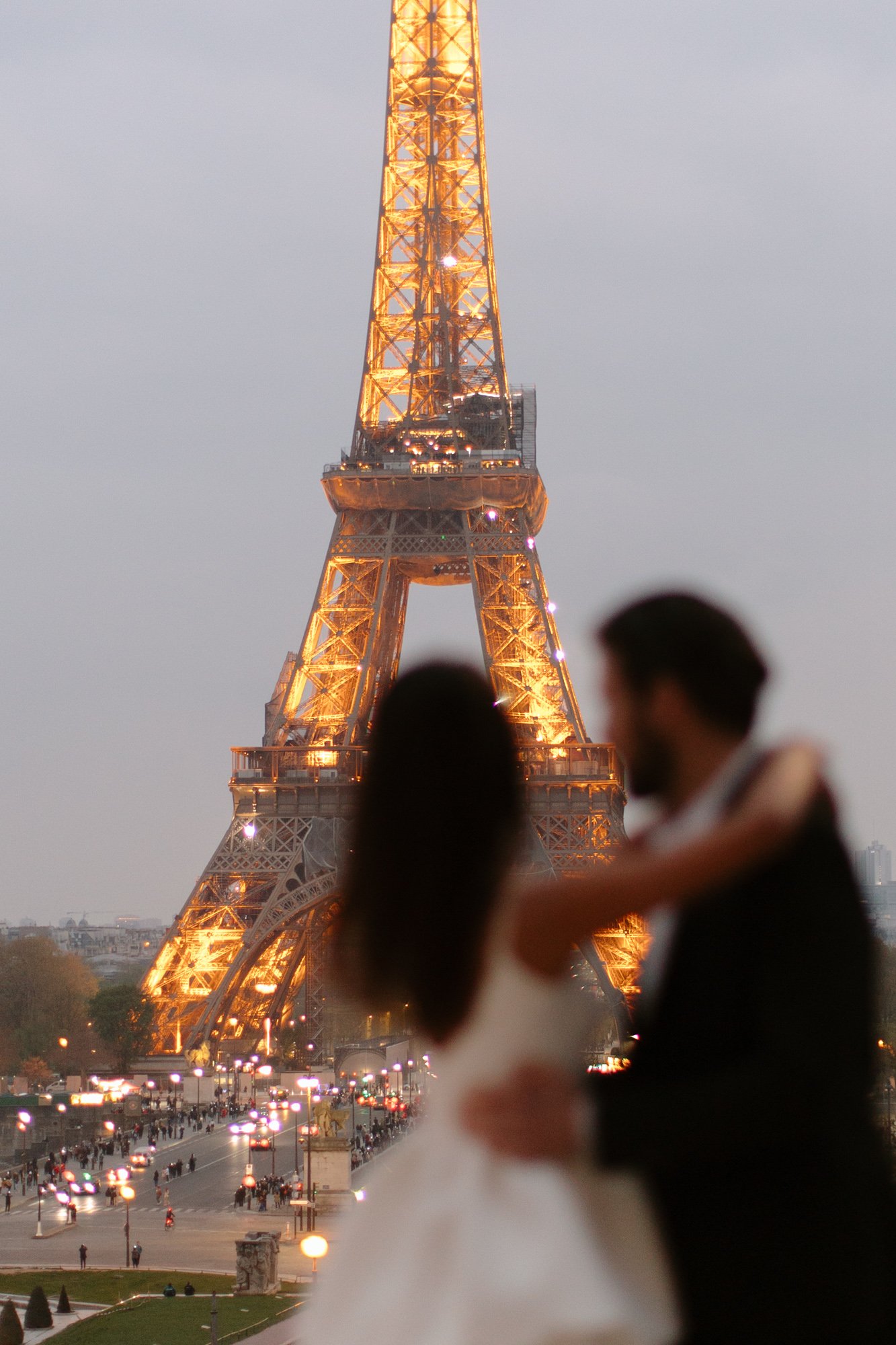 A blurred couple stands in the foreground, with the illuminated Eiffel Tower and a busy Paris street in the background at dusk.