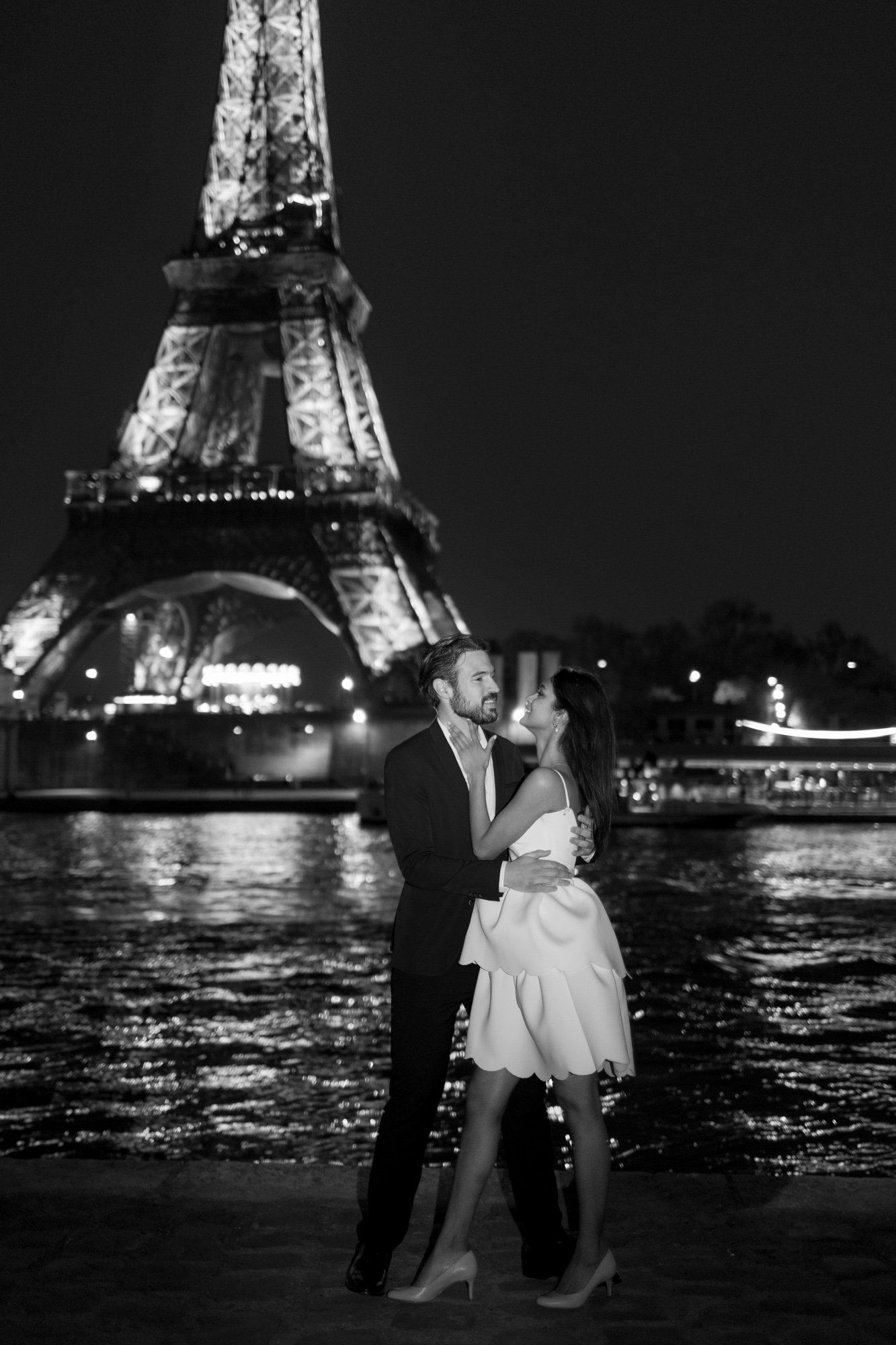 A man and woman, both dressed formally, stand smiling and embracing in front of the illuminated Eiffel Tower at night by the river. 