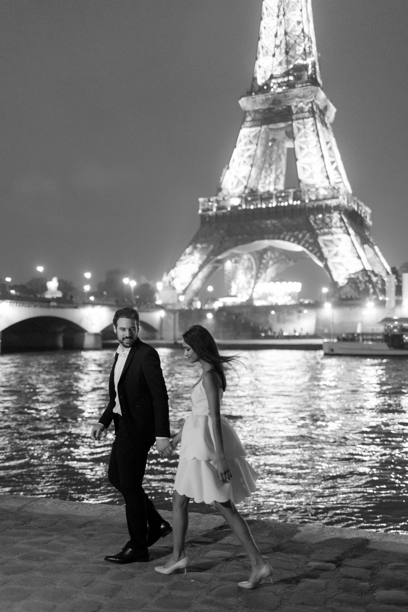 A man in a suit and a woman in a white dress walk hand in hand beside the Seine River at night, with the illuminated Eiffel Tower in the background.