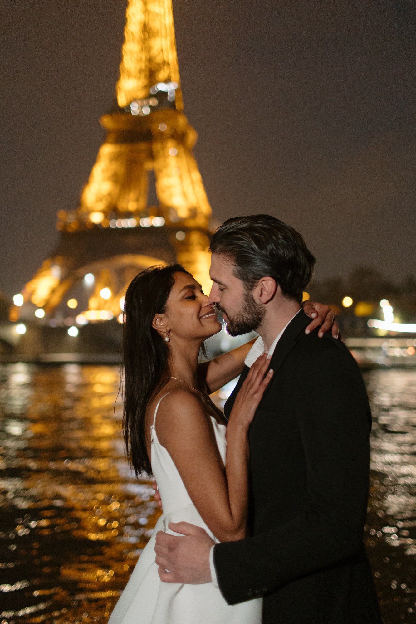 A couple in formal attire embraces and smiles at each other at night in front of the illuminated Eiffel Tower by the water. Paris Wedding Photoshoot