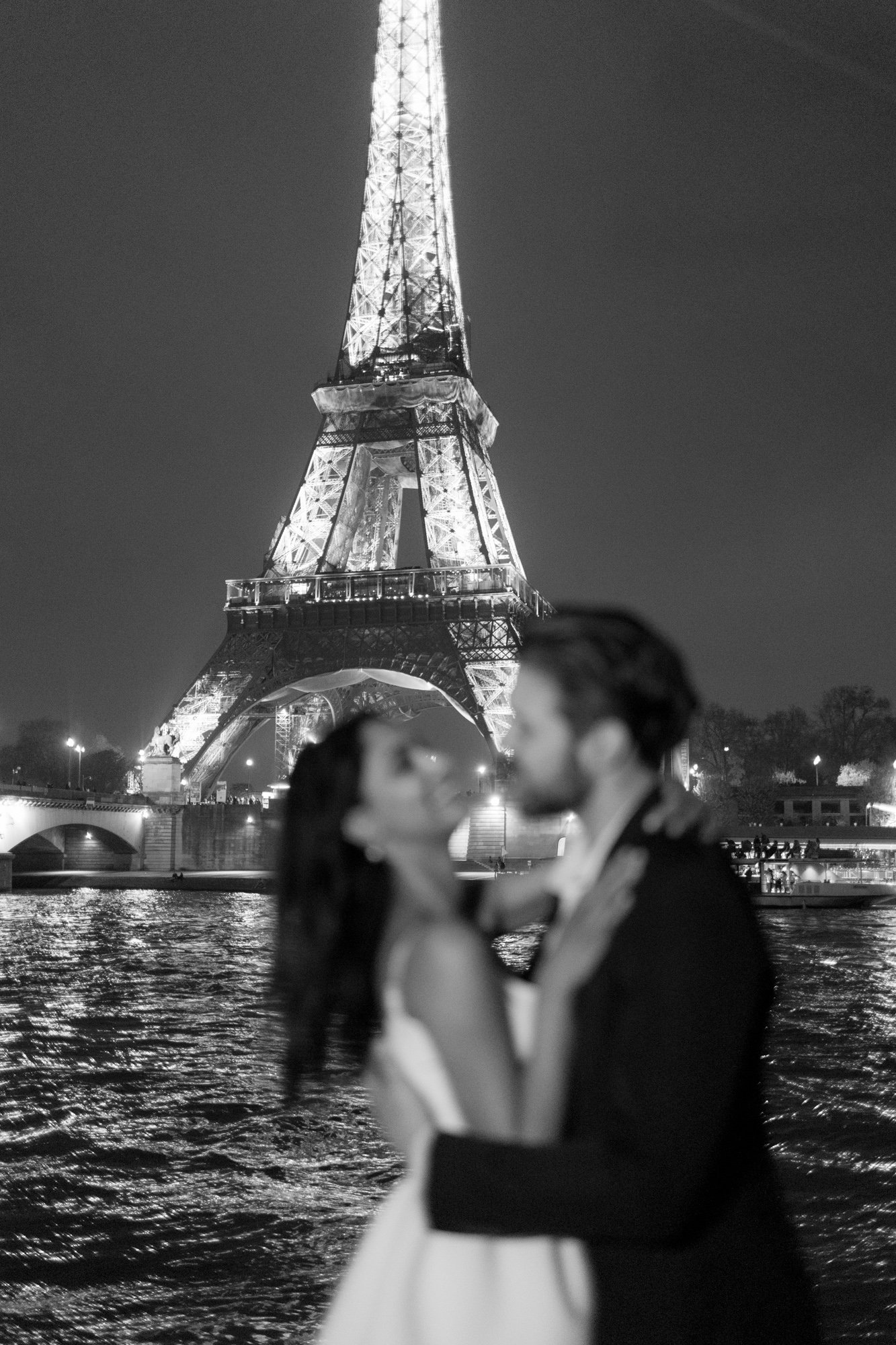 A couple stands in an embrace in front of the illuminated Eiffel Tower at night, with the river in the foreground. The couple is out of focus; the Eiffel Tower is in sharp focus.