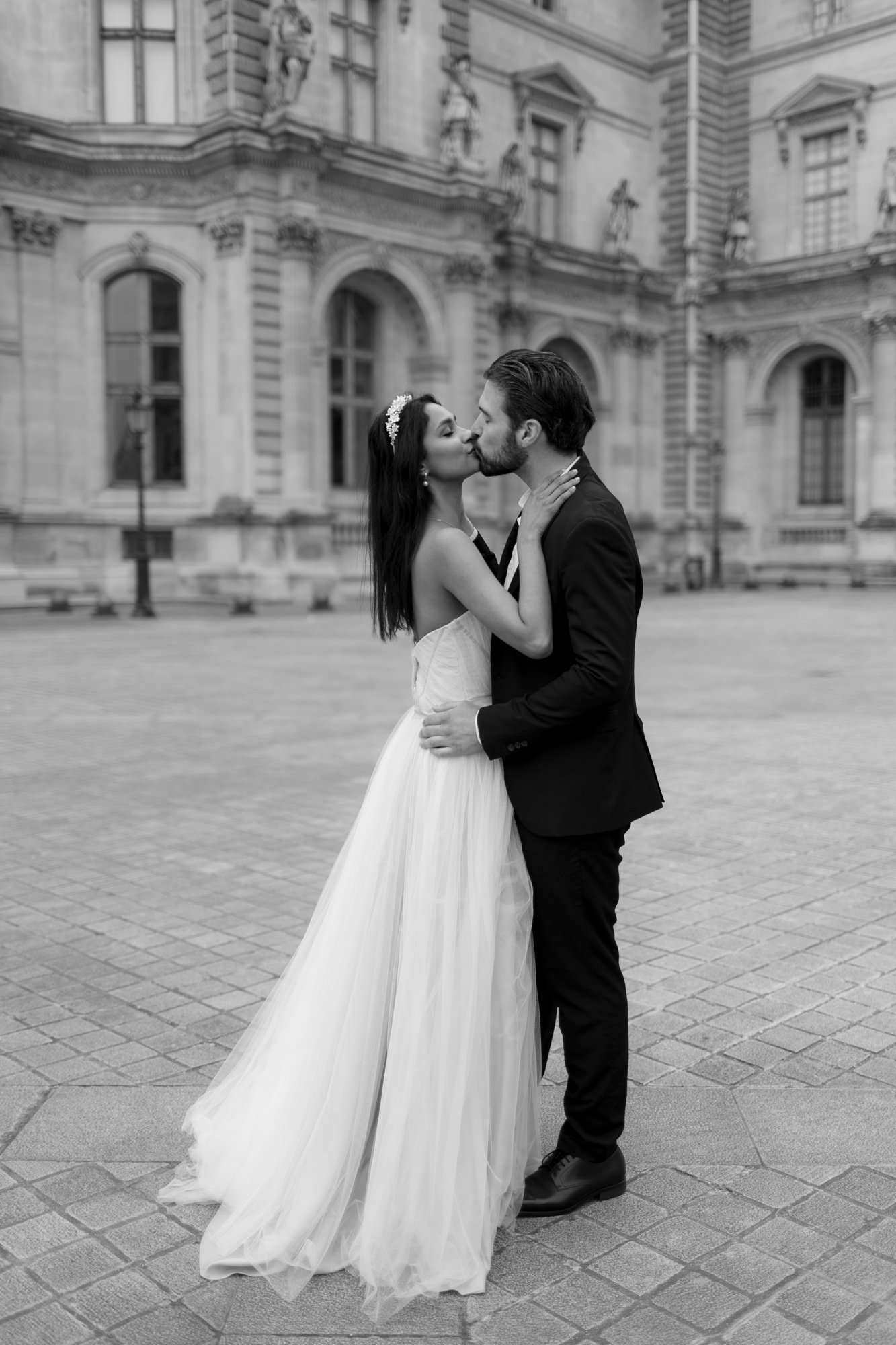 A bride and groom share a kiss while standing outside an ornate historic building, both dressed in formal wedding attire. Paris Wedding Photoshoot