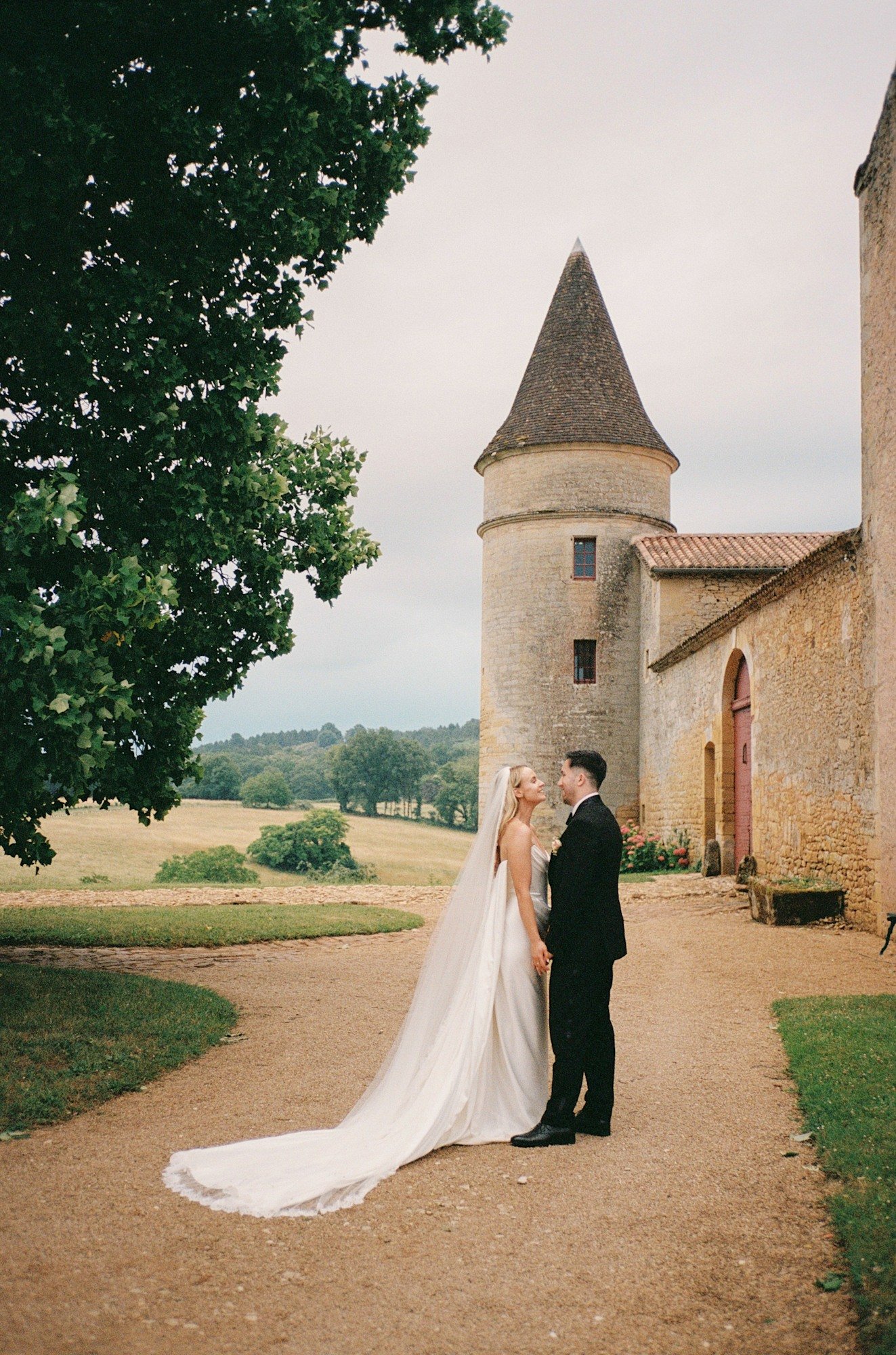 A bride and groom stand facing each other on a path outside a stone building with a turret, surrounded by greenery and fields.